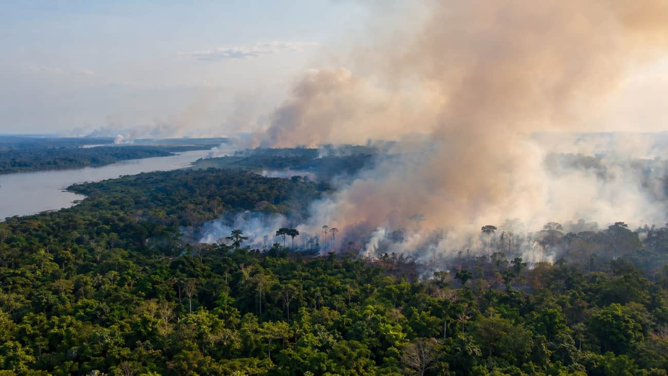 Coluna de fumaça de uma queimada subindo de uma área de floresta amazônica, com um rio ao fundo